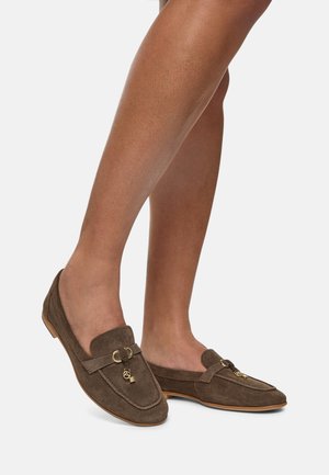 Mocassini in suede marrone con suola piatta, dotati di dettagli in metallo e di un charm decorativo sul davanti, che mostrano un design classico.