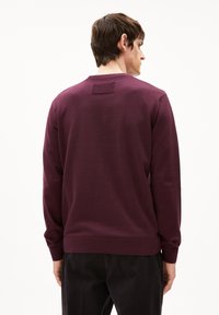 Bordeaux-roter Pullover mit Rundhalsausschnitt, langen Ärmeln und gerippten Bündchen; hat ein rechteckiges Patch auf der Rückseite und besteht aus weichem Stoff.