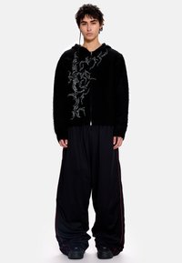 Juodas pūkuotas gobtuvinis džemperis su baltu gėlių siuvinėjimu, derintas su laisvomis juodomis kelnėmis su raudonomis juostelėmis ir derančia avalyne.