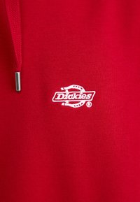 Hanorac roșu din material de bumbac, având un logo "Dickies" brodat alb, o textură catifelată și o croială standard cu sireturi.