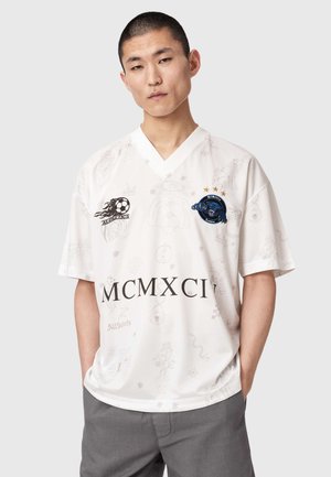 Młody mężczyzna ubrany w biały sportowy jersey z dekoltem w serek, z czarnymi rzymskimi cyframi, logo i subtelnymi tatuażowymi wzorami, ręce w kieszeniach szarych spodni.