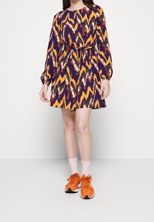 Femme portant une robe à motifs multicolores à manches longues, une ceinture à la taille, des chaussettes blanches et des baskets orange vif, debout devant un fond blanc.