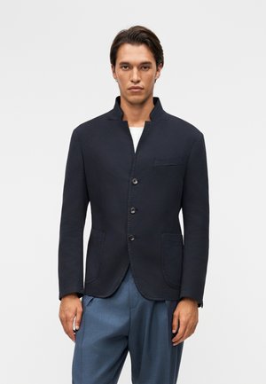 Vyras, dėvintis tamsiai mėlyną švarką su stovinčiu apykakliu, trimis sagtimis ir priekiniu kišenėmis, derinamas su mėlynomis kelnėmis.