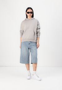 Hoodie gris avec une poche avant, associé à un short bermuda en denim bleu clair. Le mannequin porte des baskets blanches et des lunettes de soleil noires.