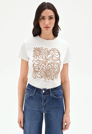 Femme aux cheveux foncés portant un T-shirt blanc à manches courtes avec un imprimé floral abstrait marron et un jean taille haute bleu, debout devant un fond blanc.