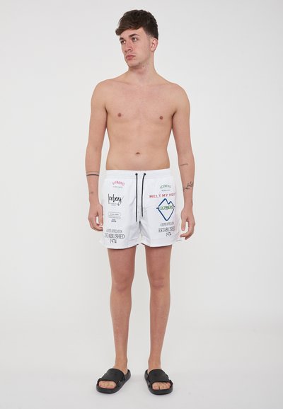 Pantaloni da nuoto bianchi con coulisse nera, caratterizzati da testo e loghi multicolori. Il modello è in piedi a piedi nudi, con la suola dei sandali visibile. Design semplice.