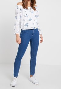 White off-the-shoulder top with blue floral print, long sleeves, and tie detail, paired with blue skinny jeans and white sneakers.