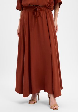 Femme portant une longue jupe orange brûlé avec une taille à cordon et des sandales à talons hauts marron clair, debout devant un fond uni.