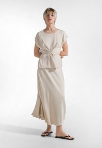 Lysebeige blød stoftop med tætsiddende talje og bindedetalje, kombineret med en lang, flydende nederdel med sideslids og elegante sorte sandaler.