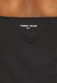 Black ribbed tank top featuring a V-shaped neckline. The brand "TOMMY JEANS" is embroidered in white on the back with a color accent.