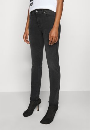 Personne portant un jean noir ajusté, des bottines chaussettes à talons noires et une chemise blanche rentrée, debout devant un fond clair uni.