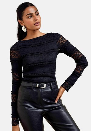 Vrouw draagt een zwarte kanten top met lange mouwen met horizontale ruches en zwarte leren broek met een zwarte riem met zilveren gesp.