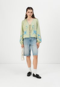 Ljusgrön blus med blå broderi, scallopad nederkant och puffade ärmar, i kombination med denim bermudashorts. Accessoarer inkluderar en vit väska.