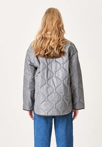 Polstret grå quiltet jakke med diamantmønster, afslappet pasform, sort kant og brede ærmer, båret over blå denimbukser.