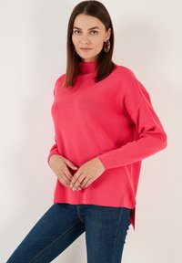 Pull en tricot rose avec un col montant, des manches longues et des fentes sur les côtés, associé à un jean bleu foncé. Texture lisse et coupe décontractée.