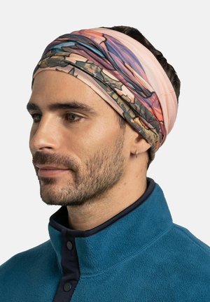 Hombre con barba corta que lleva una cinta para la cabeza de colores y un chaqueta de forro polar azul, mirando hacia la izquierda con expresión neutral sobre fondo claro.