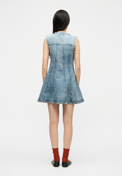 Γυναίκα που φορά ένα αμάνικο ανοιχτό μπλε τζιν φόρεμα με λεπτομέρειες με καρφιά, κόκκινες κάλτσες και μαύρα παπούτσια, στέκεται γυρισμένη με την πλάτη σε λευκό φόντο.