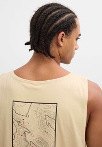 Joven con el cabello trenzado que lleva una camiseta sin mangas beige con un diseño de mapa topográfico negro y rojo en la espalda, sobre un fondo liso.