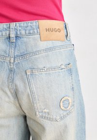 Jean en denim bleu clair avec une poche arrière légèrement usée et une étiquette beige indiquant « HUGO », porté avec un haut rose vif.