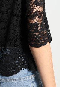 Top in pizzo nero con motivo floreale, maniche a scallop e orlo ondulato. Indossato sopra dei jeans, mostrando dettagli intricati nella texture e nel design.