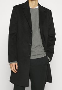 Schwarzer Wollmantel mit maßgeschneidertem Design, einreihig, mit fünf Knöpfen und Reverskragen. Über einem grauen Pullover getragen.