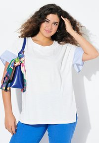 Valkoinen t-paita, jossa on siniset raidalliset lyhyet hihat, yhdistettynä sinisiin housuihin. Kirkkaan värinen kuvioitu huivi on sidottu siniseen käsilaukkuun.