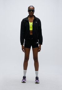 Must tuulejakk, neoonkollane lühike top, mustad lühikesed püksid valgete detailidega, lillad spordijalatsid ja tumedad päikeseprillid. Modell seisab näoga ettepoole.