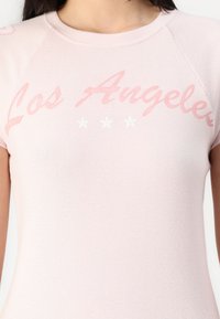 Rosa kortärmad skjorta med ”Los Angeles” i kursiv stil och tre vita stjärnor tryckta nedan. Slät textur med klassisk rund hals.