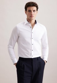 Camicia bianca con colletto strutturato, dettagli decorativi blu all'interno e bottoni scuri, abbinata a pantaloni blu navy.