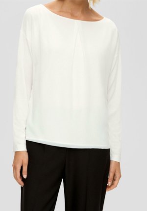 Vrouw draagt een witte blouse met lange mouwen en een plooidetail aan de voorkant, gecombineerd met een zwarte broek, staand tegen een effen achtergrond.