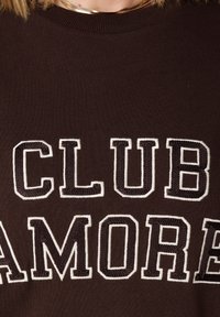 Bruine gebreide trui met grote witte geborduurde letters met de tekst "CLUB AMORE." De halslijn is geribbeld en biedt een gestructureerde afwerking.