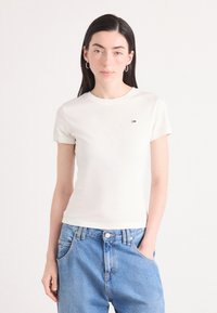 T-shirt in cotone color avorio, con collo a giro e maniche corte, e un piccolo logo blu e rosso sul petto. Indossato con jeans azzurri chiari.