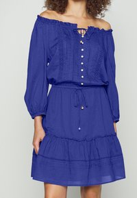 Blå off-the-shoulder klänning i lätt tyg. Har framknappar, midja med dragsko och volangkant med dekorativa mönster.