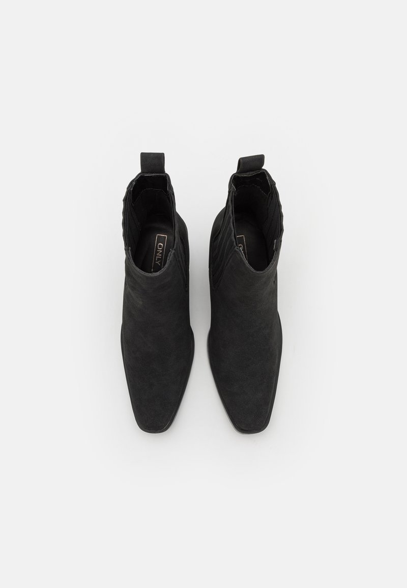 Bottes noires à cheville avec une texture en daim lisse, un bout pointu et une languette de tirage. Présentent des nervures verticales sur les côtés pour un détail de design.