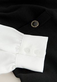 Must musternahk, millel on valgest satiinist varrukas. Omab volangiääre ja kahte läbipaistvat nööpi, mille rõhutamiseks on must nööp kuldse äärisega.