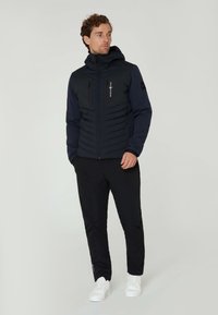Mörkblå isolerad jacka med dragkedja fram, huva och två bröstfickor; i kombination med svarta byxor och vita sneakers.