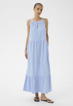 Donna in piedi che indossa un vestito lungo azzurro senza maniche con scollo arricciato e tasche laterali, abbinato a infradito nere.