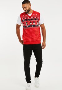 Rode gebreide mouwloze trui met een gebreide halslijn met sneeuwvlokken en kabouters, gedragen over een wit T-shirt, gecombineerd met zwarte broek en witte sneakers.