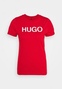�Červené bavlněné tričko s kulatým výstřihem, krátkými rukávy a výrazným bílým logem "HUGO" na hrudi. Hladký materiál, standardní střih.