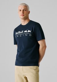 Junger Mann mit kurzen blonden Haaren, der ein dunkelblaues Red Bull Racing T-Shirt und beige Hosen trägt, vor einem schlichten grauen Hintergrund steht.