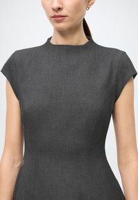 Grå tætsiddende kjole med korte kapærmer, høj halsudskæring og tekstureret stof. Sømme detaljer fremhæver den strukturerede silhuet.