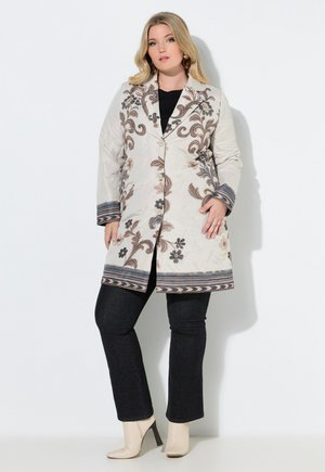 Nainen, joka pukeutunut kermanväriseen kukkakuosilliseen takkiin mustien housujen ja kermanväristen korkokenkien kanssa, seisoo valkoista taustaa vasten.