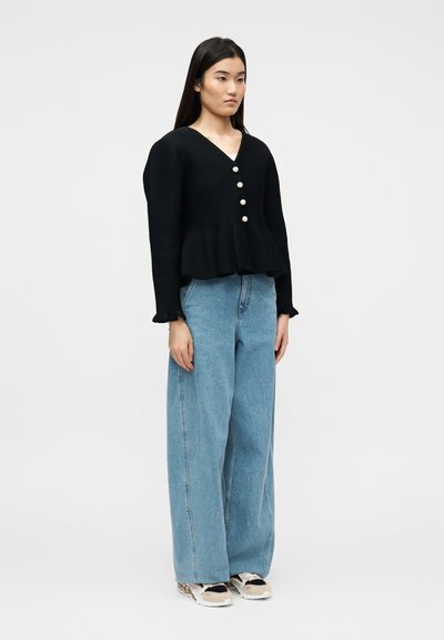 Νεαρή γυναίκα στέκεται φορώντας μαύρο πουλόβερ με κουμπιά και πεπλουμ, φαρδιά ανοιχτό μπλε τζιν παντελόνια και μπεζ με μαύρα αθλητικά παπούτσια, σε απλό ανοιχτό φόντο.