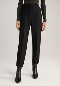 Pantalon noir ajusté avec une coupe droite, présentant des plis avant et une taille avec fermeture à bouton. Porté avec des bottines noires élégantes.