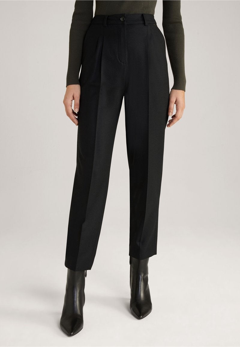 Pantalon noir ajusté avec une coupe droite, présentant des plis avant et une taille avec fermeture à bouton. Porté avec des bottines noires élégantes.