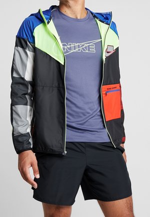 Homme portant un short noir, un t-shirt bleu Nike et une veste zippée colorée avec des panneaux bleus, vert néon, gris, noir et orange.