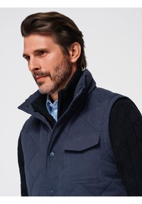 Gilet trapuntato blu navy con colletto alto, dotato di tasca frontale e chiusura a bottoni, indossato sopra un maglione nero a maglia e una camicia azzurro chiaro.