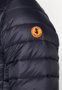 Granatowa puchowa kurtka o pionowym wzorze pikowania, z pomarańczowym okrągłym logo z napisem "SAVE THE DUCK" na lewym ramieniu.