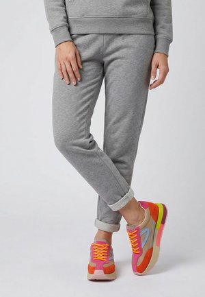 Person iført grå joggingbukser og farverige sneakers med orange snørebånd står med det ene ben krydset over det andet.