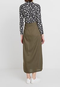 Langer olivgrüner Rock aus leichtem Stoff, kombiniert mit einem langärmeligen schwarzen Hemd mit weißem Blumenmuster. Weiße Turnschuhe getragen.